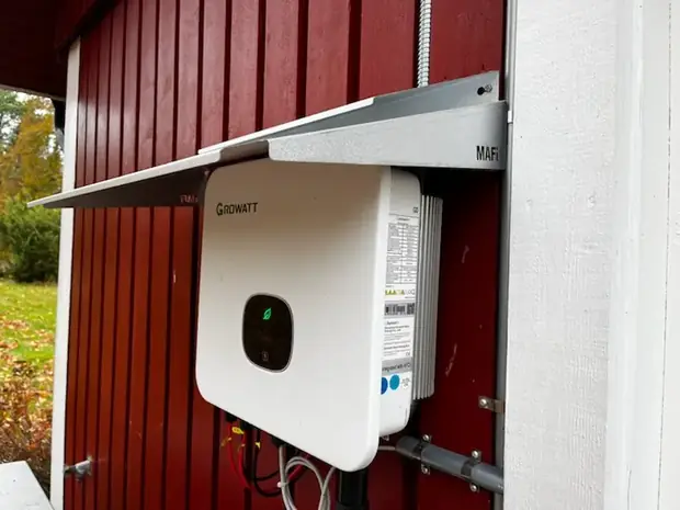 Vit växelriktare monterad på en röd träfasad under ett skyddande takutsprång.